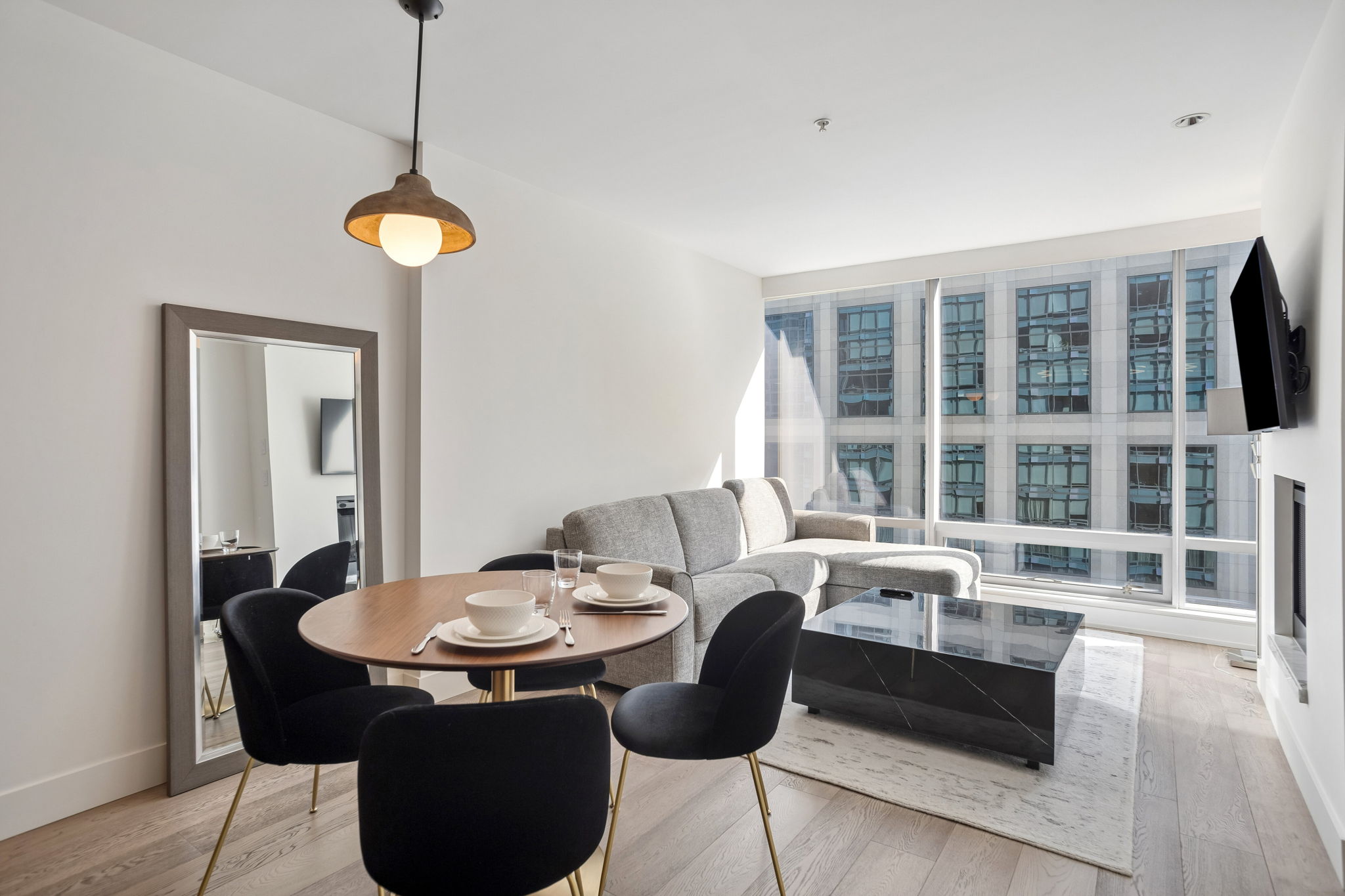 The living and dining room seen from the entry, with a tall leaning mirror and city towers through the windows.