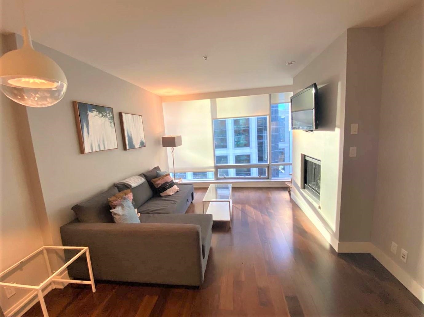 The living room, a grey sofa on dark hardwood with the window wall and a gas fireplace along the right.