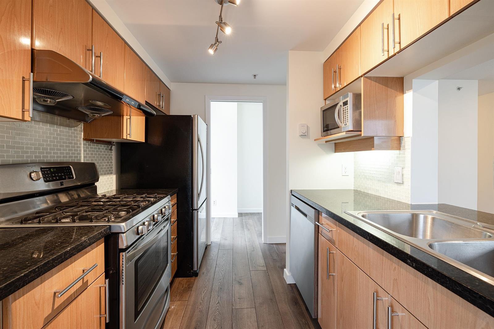 The kitchen along its length: a gas range under a stainless hood, tiled splashback, double sink and dishwasher on dark stone counters.