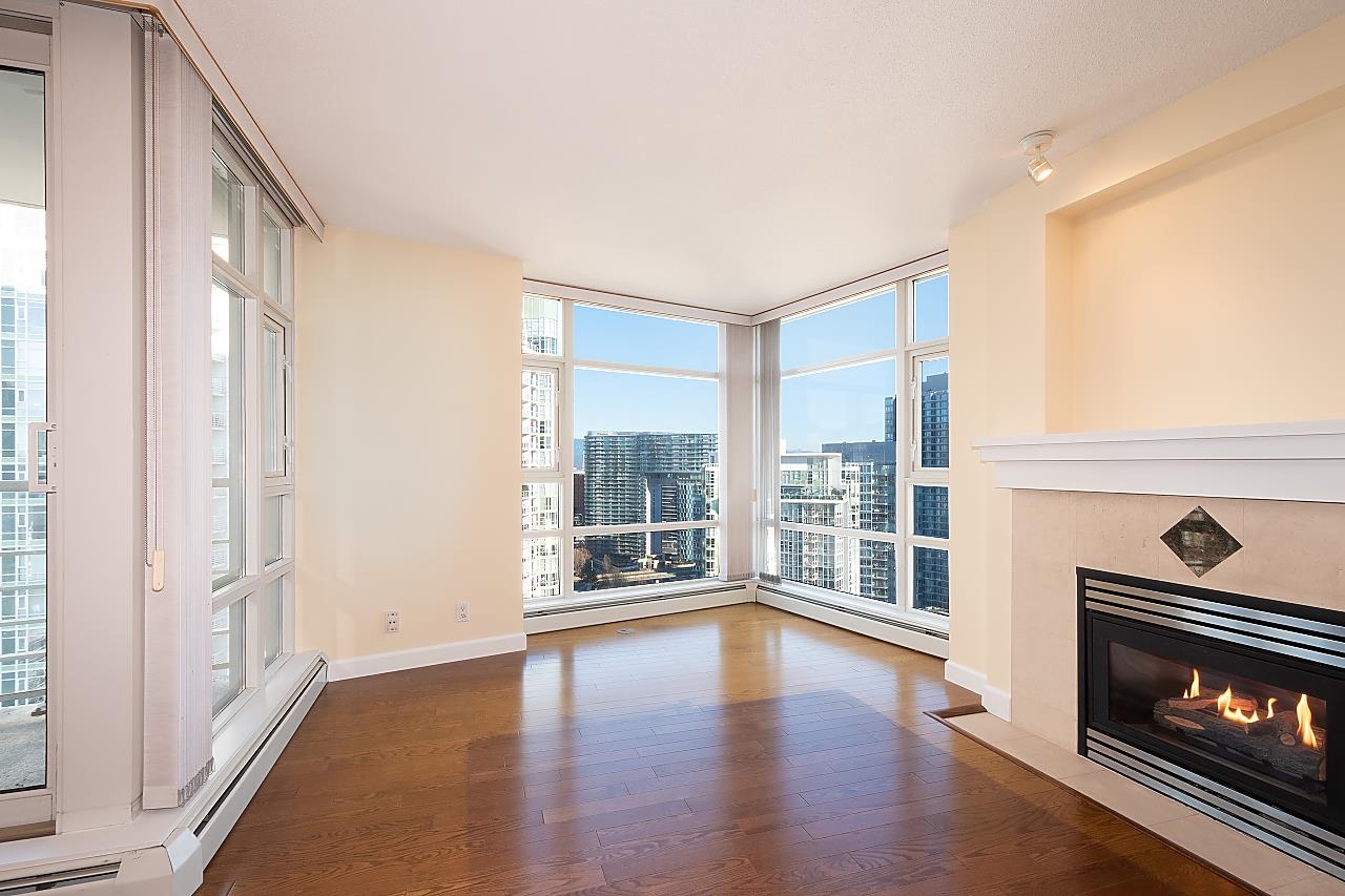 The living room from the dining end, windows turning the corner and the fireplace alight at the right.