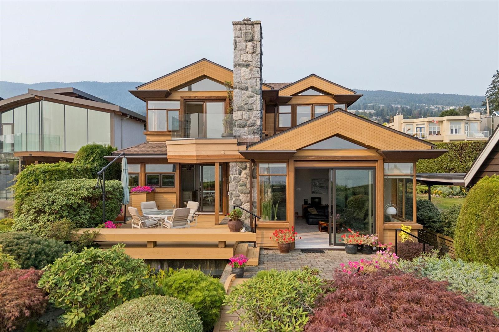 The house from the garden, the stone chimney running up through timber and glass, shrubs and maples below.