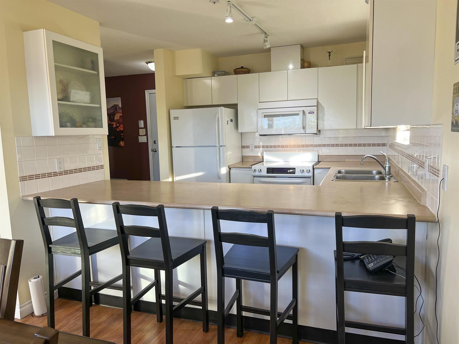 The kitchen, white cupboards around a breakfast bar with stools along one side.