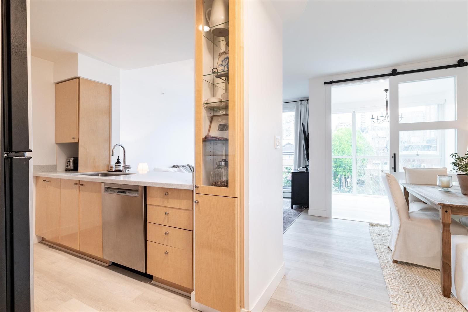The entry looking through, maple cabinets and a glass-fronted cabinet on the left, the dining area and balcony door at the far end.