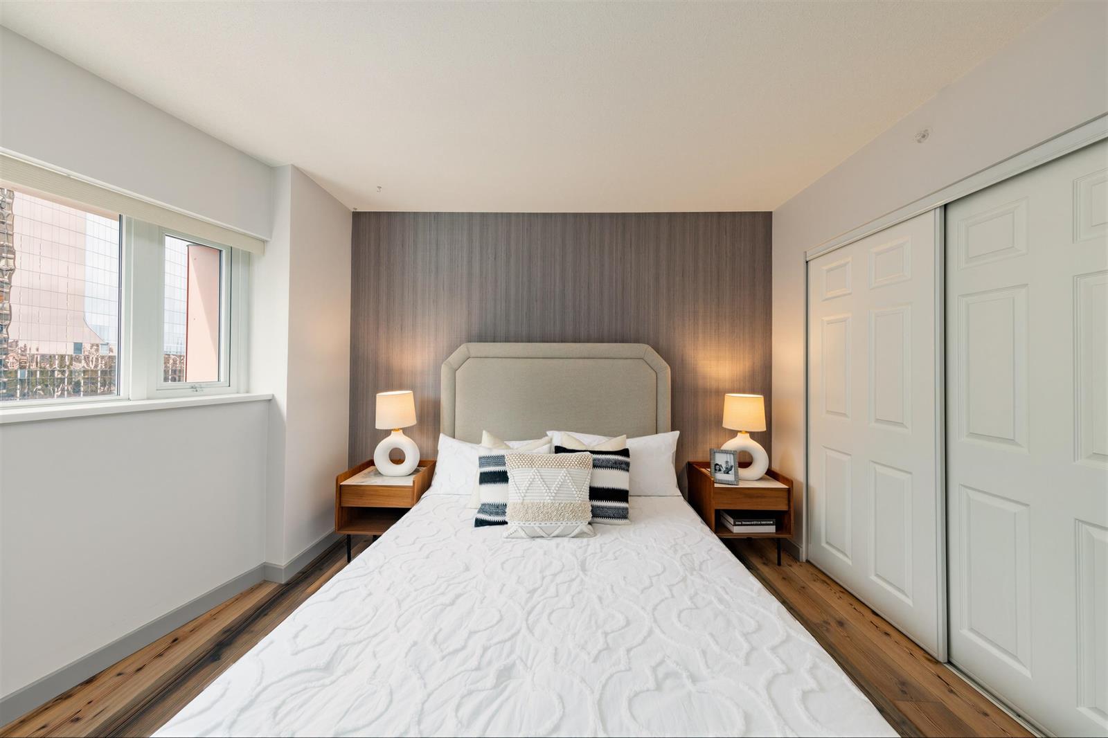 A bedroom, the bed between two lamps with the window at its side and the closets opposite.