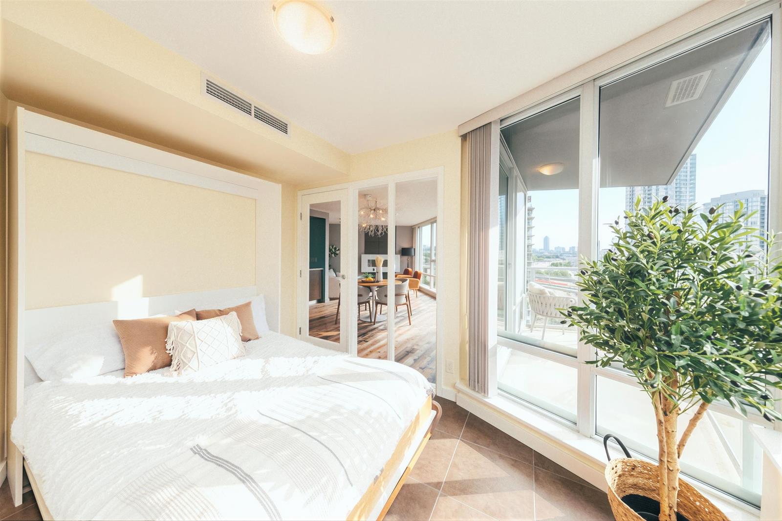 A bedroom looking out to the living room, the bed at the window with a plant beside it.