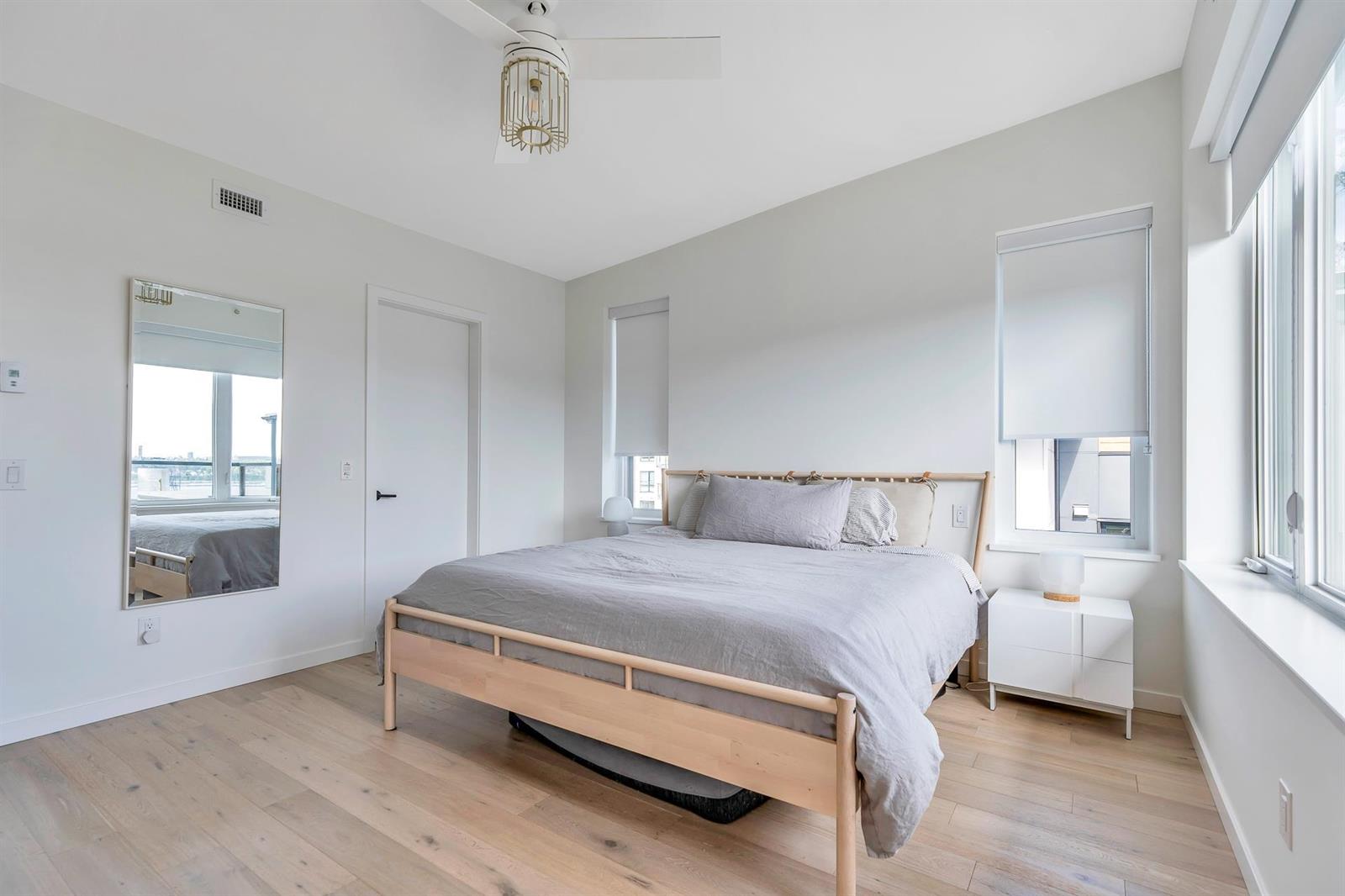 A bedroom, bright, with the bed against the far wall and windows to one side.