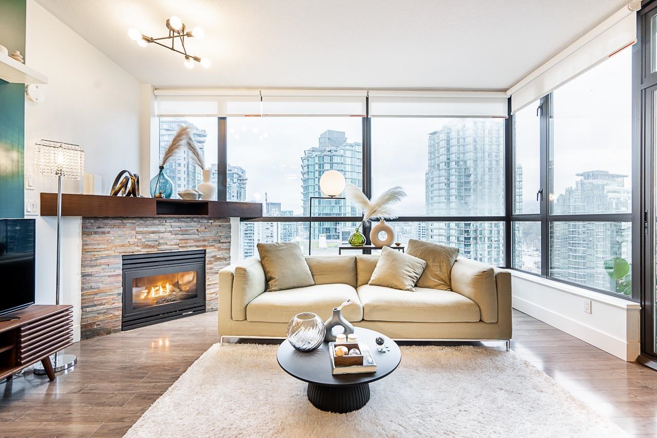 The living room turning the corner in glass, a gas fireplace on the left and the towers of Coal Harbour beyond the windows.