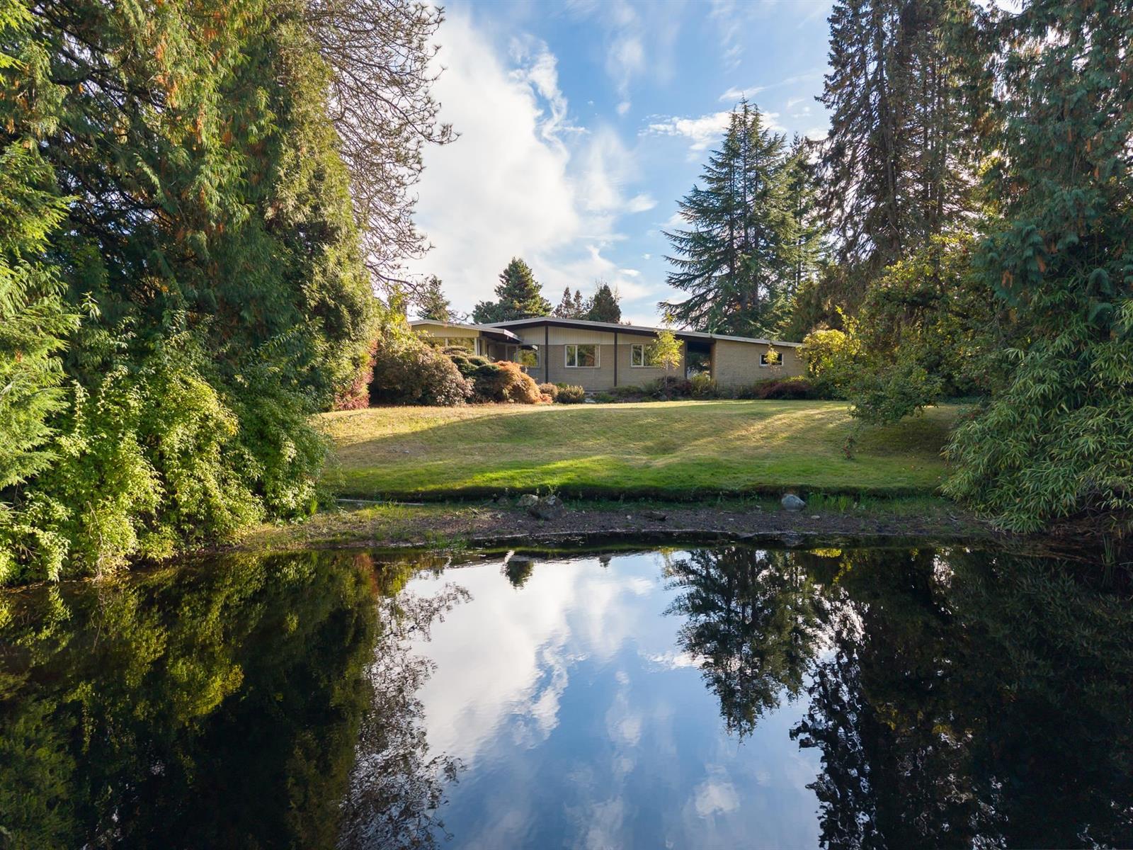 The house seen across Elveden Lake, low and long behind the lawn, with the sky and cedars reflected in the water.