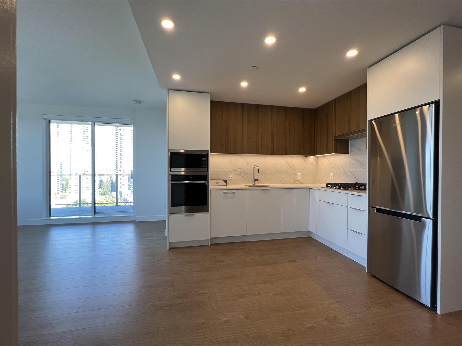 The kitchen and living room as one space, cupboards along the far wall and windows running down the side.