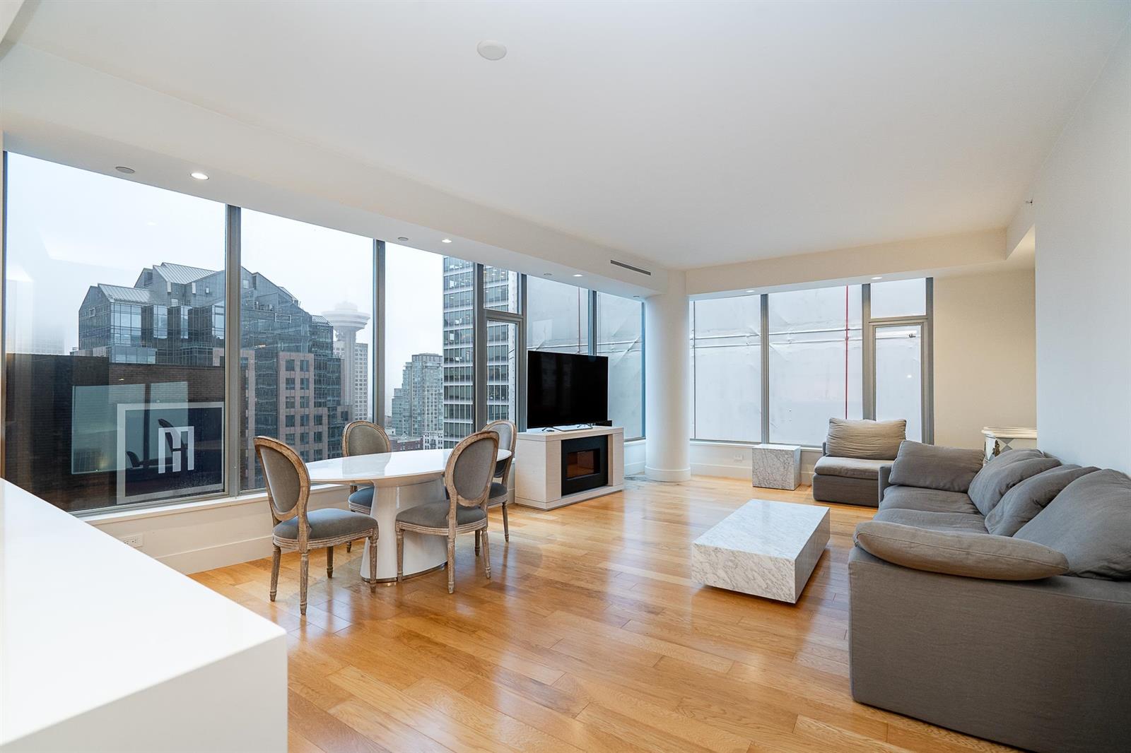 The living room, a wall of glass along two sides with the dining table at the far end and the city outside.