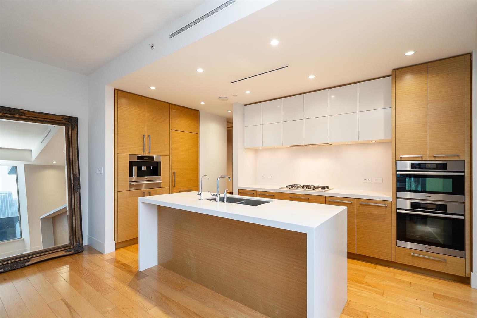 The kitchen, a long island with a sink facing a run of pale cabinetry, wall ovens and a gas cooktop.