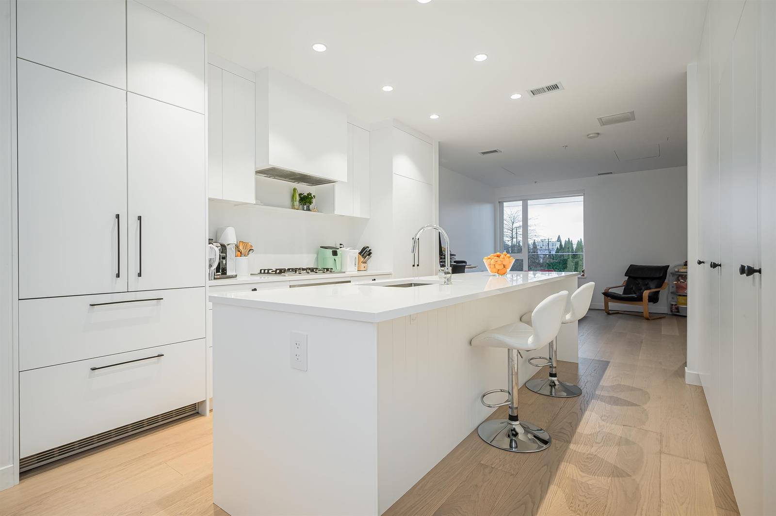 The kitchen: a long white island with two stools, cabinetry to the ceiling, and the floor running on past it to the living end and its window.