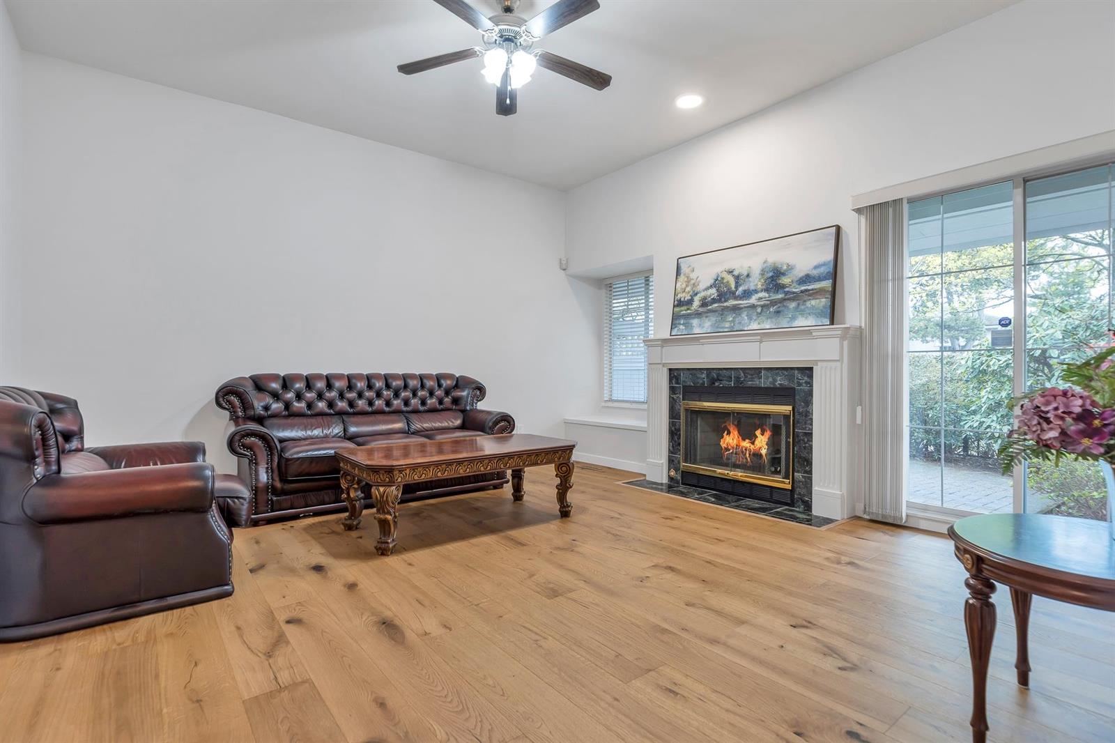 The living room, a gas fireplace lit under its mantel between a window and the sliding door to the patio, on wide oak boards.