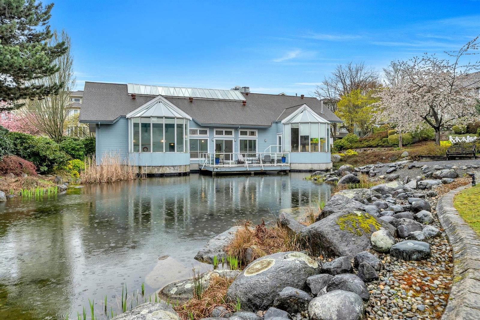 The complex across its pond — pale blue houses with conservatory windows reflected in the water, blossom on the far bank.