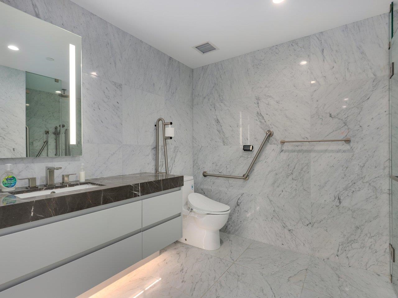 The bathroom, tiled floor to ceiling in white marble, with a wall-hung vanity, backlit mirror and grab rails.